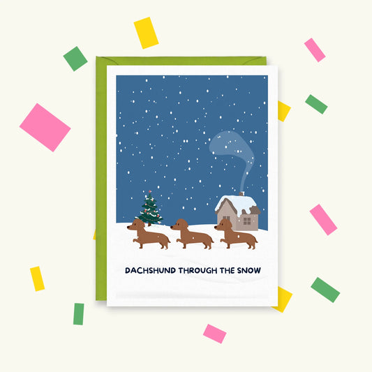 Greeting card of three dachshunds walking through snow past a Christmas tree and cottage, text "Dachshund Through The Snow"