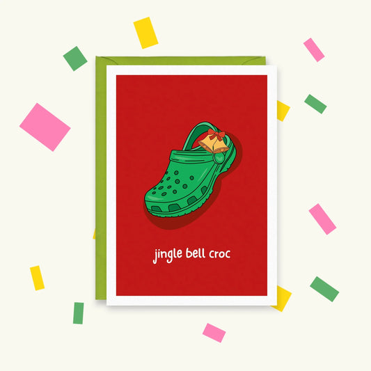 Greeting card with a green Croc clog wearing a golden jingle bell charm on a red background, reading "jingle bell croc"